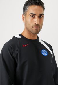 Sudadera negra con el logo rojo de Nike, acento blanco en el hombro y emblema azul del Paris Saint-Germain. Tejido ligero y suave, cuello redondo.