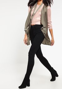 Ljus olivgrön väst över en rosa stickad tröja, parat med svarta skinny jeans och svarta ankelskor. Släta texturer och lagerdesign.