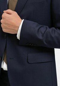 Marineblå skräddarsydd kostymjacka med spetsig slag, enkel ficka och knappdetaljer på ärmarna. Vit skjorta synlig under.