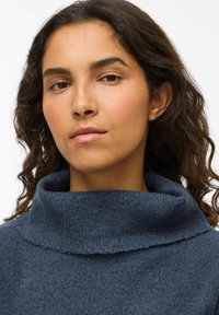 Un pull texturé bleu foncé avec un col haut et large. Le tissu semble doux et confortable, arborant un design minimaliste.
