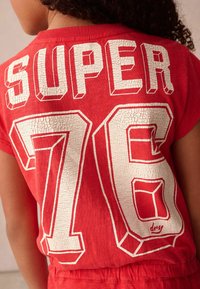 Röd T-shirt med kort ärm som visar "SUPER" i vit, sprucken text ovanför "76" i en fet, omrisstad stil. Mjuk, avslappnad tygkänsla.