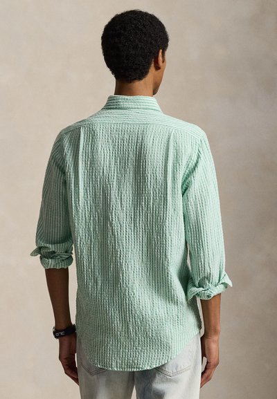 Egy férfi háttal, világoszöld, texturált, hosszú ujjú inget visel feltűrt ujjakkal és világos színű nadrágot semleges háttér előtt.