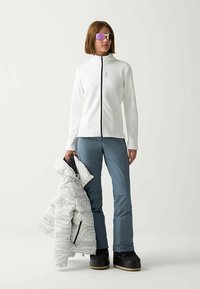 Fehér cipzáras dzseki fekete díszítéssel, szürke mintás pufi dzseki, kék sí nadrág és fekete hócipő, amelyet egy álló alak tart.