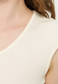 Κοντινό πλάνο ενός ατόμου που φοράει αμάνικο, εκρού ριπ τοπ με στρογγυλεμένο λαιμό και εμφανή ανοιχτό καφέ μαλλιά.