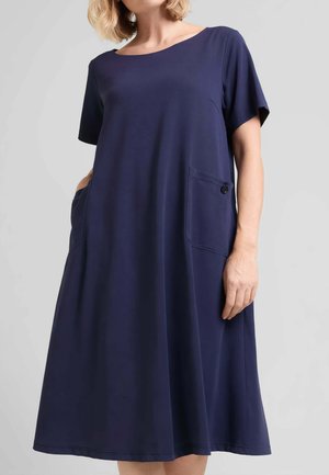 Donna che indossa un vestito blu navy ampio, lunghezza al ginocchio, con maniche corte e una tasca frontale con bottone su uno sfondo semplice.