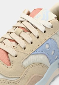 Beige sneaker with pastel blue and pink accents, textured fabric, and cream laces. Features a distinct logo and a wavy design along the sides.