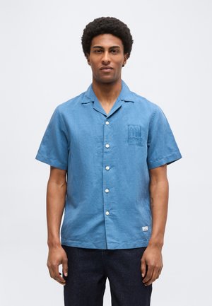 Homme aux cheveux courts et bouclés portant une chemise bleue à manches courtes boutonnée et un pantalon foncé, debout devant un fond blanc uni.