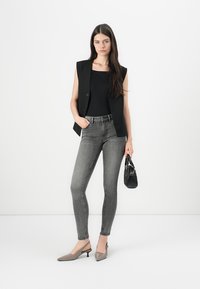 Black sleeveless blazer over black top, paired with fitted gray jeans and gray slingback heels. Holds a small black handbag.