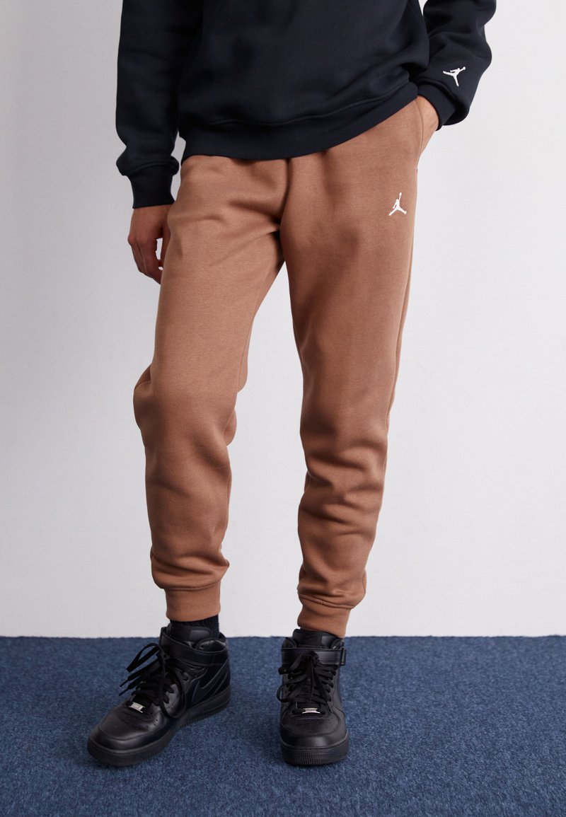 Brown sweatpants with elastic cuffs feature a white logo on the left thigh, paired with black high-top sneakers on a blue carpet.