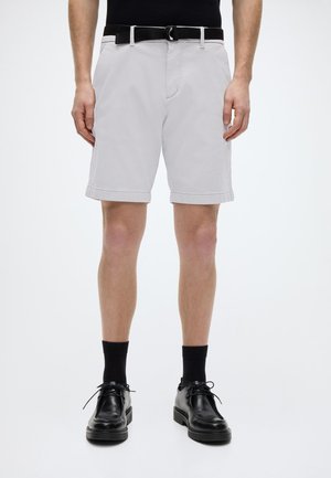 Shorts gris clair avec un motif texturé, revers roulés et ceinture noire. Associés à des chaussures et des chaussettes noires pour un look harmonieux.