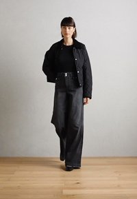 Prošívaná černá bunda, upnutý černý top a tmavé džíny s vysokým pasem a širokými nohavicemi. Modelka stojí na dřevěné podlaze před jednobarevným pozadím.