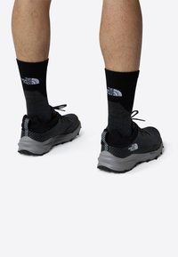 Zapatillas deportivas negras con suelas grises, que presentan un upper texturizado y acentos blancos. Complementadas con calcetines oscuros que muestran un logo.