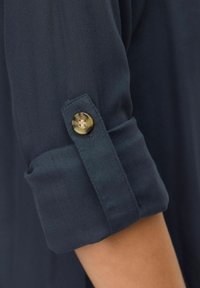 Manche de chemise boutonnée bleu marine foncé, en tissu doux, avec un poignet retroussé et un détail de bouton doré pour un maintien sécurisé.