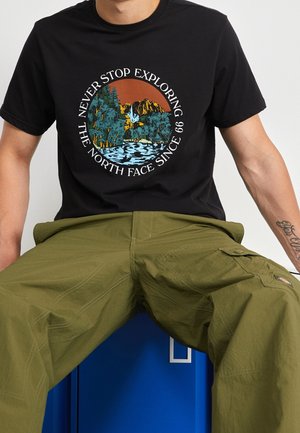 Homme portant un t-shirt noir avec un graphisme circulaire représentant la nature et un texte, associé à un pantalon cargo vert olive, assis sur un casier bleu.