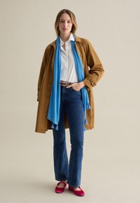 Brauner langer Mantel, weißes Hemd, blauer Schal, dunkelblaue Schlagjeans und pinke Ballerinas. Der Mantel hat Knöpfe; der Schal hat Fransen.