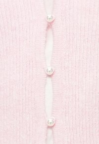 Ροζ πλεκτή ζακέτα με μαργαριταρένια κουμπιά στο εμπρός μέρος, με υφή και λευκή εσωτερική στρώση ορατή στο άνοιγμα.