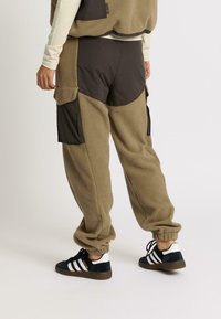 Personne portant un pantalon cargo en polaire beige avec des patchs noirs et des poignets élastiques, associé à des baskets noires et blanches Adidas.