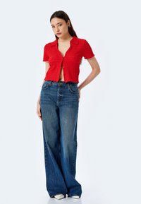 Camicia rossa a maniche corte con texture e bottoni frontali abbinata a jeans blu a vita alta e a gamba larga. Il modello è in piedi davanti a uno sfondo neutro.