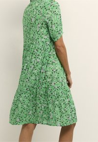 Robe fleurie verte à manches courtes, taille froncée et ourlet évasé. Présente des motifs de fleurs blanches et noires sur un tissu léger.