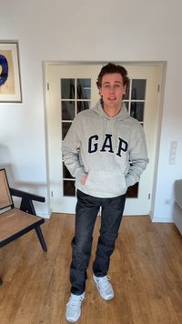 Young man standing indoors wearing a gray GAP hoodie, dark pants, and white sneakers, with hands in pockets and a neutral expression.