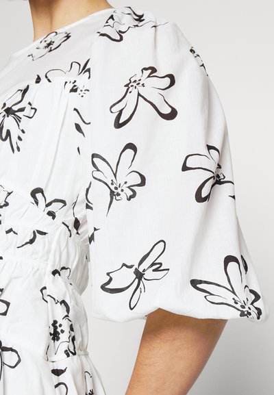 Λευκή μπλούζα με μαύρα αφηρημένα λουλουδάτα σχέδια, φουσκωτό κοντό μανίκι και συγκεντρωμένες ελαστικές λεπτομέρειες στη μέση και το στήθος.