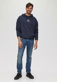 Marineblauwe hoodie met voorzak en trekkoord, gecombineerd met lichtblauwe jeans en zwarte sneakers, staand op een neutrale achtergrond.