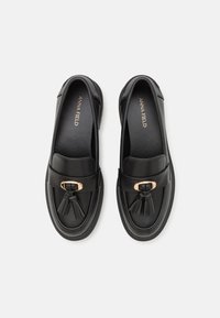 Mustad nahast mustad loafers ümmarguse varbaga, millel on kuldse tooniga pandla ja esiosas on tõmblukuga detail. Sile tekstuur ja minimaalne disain.