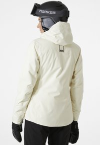 Witte waterdichte skijas met een capuchon, voorzien van een subtiel logo op de achterkant, gecombineerd met zwarte handschoenen, tegen een effen achtergrond.