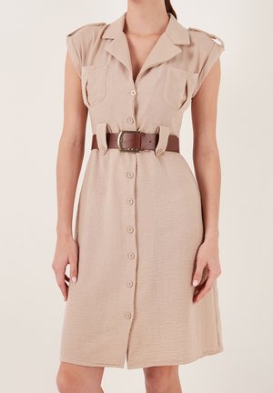 Femme portant une robe beige sans manches boutonnée avec des poches poitrine et une ceinture marron à la taille, debout devant un fond uni.