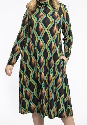 Pitkähihainen, polvipituinen mekko, jossa on vihreä, ruskea, musta ja sinivihreä geometrinen timantinmuotoinen kuvio, ihmisen yllä, jolla on käsi taskussa.