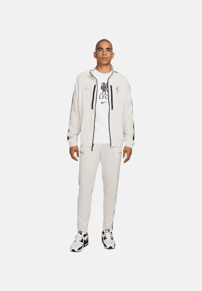 Un chándal gris claro con detalles en negro; chaqueta con cremallera, camiseta blanca con el logo de Liverpool y pantalones de chándal a juego. Zapatillas negras y blancas.