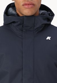 Chaqueta con capucha en color azul marino que cuenta con un exterior suave, cierre de cremallera y un pequeño logo blanco en el pecho. Forro interior suave visible en la capucha.