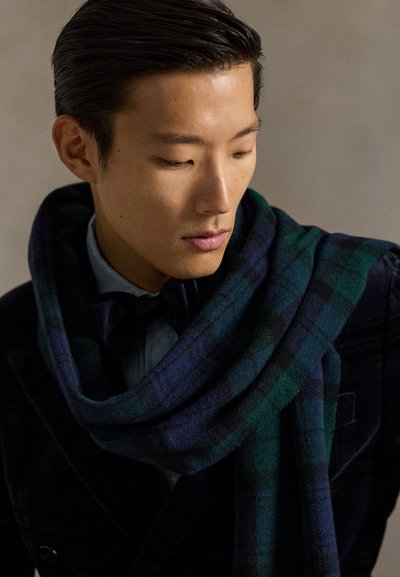 Cachecol de lã aos quadrados em azul-marinho e verde, colocado sobre um casaco escuro. Apresenta uma textura macia e um design amplo e fluido, complementando o conjunto.