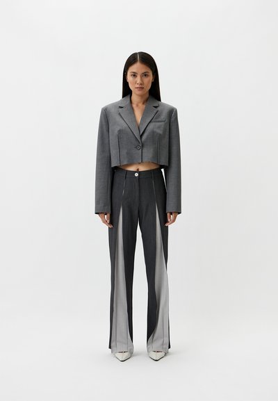 Gray cropped blazer with a single button, paired with high-waisted gray pants featuring sheer panels and split hems, and pointed white heels.