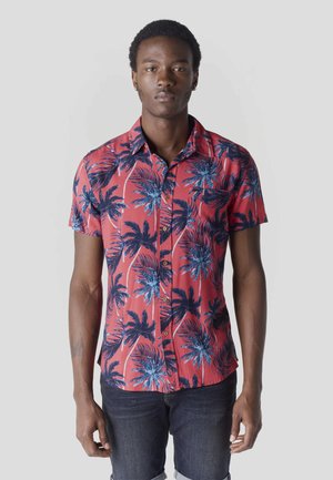 Joven de pie mirando hacia adelante, vistiendo una camisa roja de manga corta con botones y estampados de palmeras azules, y jeans de mezclilla oscura.