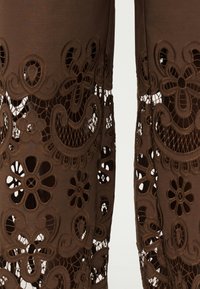 Καφέ ύφασμα με λουλουδάτα και πασλέ δαντελωτά μοτίβα με κοψίματα, που διαθέτει περίπλοκη ραφή και λεπτομέρειες από δίχτυ στα μπατζάκια του ρούχου.