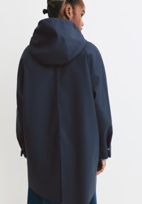 Marineblå vandtæt jakke med hætte, der har et lige design, løs pasform og knapærmer. Rene linjer og glat overflade.