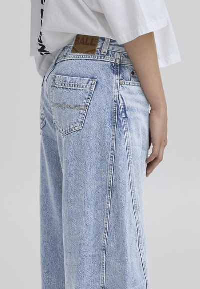 Svijetloplave široke traperice s visokim strukom i kontrastnim šavovima. Ima bočne džepove i brendiranu smeđu kožnu etiku na struku.