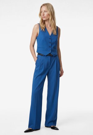 Femme debout portant un gilet bleu sans manches boutonné et un pantalon large assorti, avec des chaussures noires à bout pointu, sur un fond blanc.