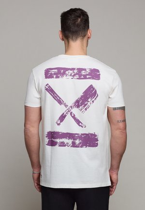 Mann mit kurzen dunklen Haaren, der ein weißes T-Shirt mit lila verwaschenem Motiv von gekreuzten Fleischbeilen auf dem Rücken trägt, steht vor grauem Hintergrund.