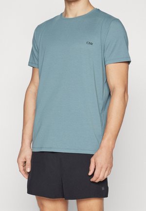 Mann trägt ein gedämpftes blaues T-Shirt mit kurzen Ärmeln und einem kleinen schwarzen Logo auf der Brust sowie schwarze Sportshorts. Er steht vor einem weißen Hintergrund.