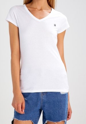 Mujer vistiendo una camiseta blanca ajustada con cuello en V y shorts de mezclilla azul desgastados, de pie con los brazos relajados a los lados.