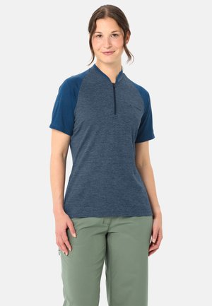 Jonge vrouw in een blauw shirt met korte mouwen en rits, en lichtgroene broek, staand tegen een effen lichte achtergrond.