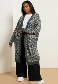 Grå mönstrad cardigan med öppen front, med leopardmönster, ribbade ärmslut och fåll, bärs över en vit t-shirt och svarta byxor.