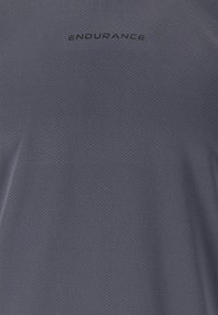 Harmaa urheilupaita, joka on valmistettu teksturoidusta kankaasta. Rintapielessä mustalla tekstillä sana "KESTÄVYYS". Lyhyet hihat ja rento malli.