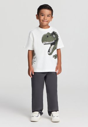 Biała koszulka z grafiką zielonego dinozaura, w zestawieniu z ciemnoszarymi spodniami i białymi trampkami z czarnymi akcentami. Casualowy design dla dzieci.