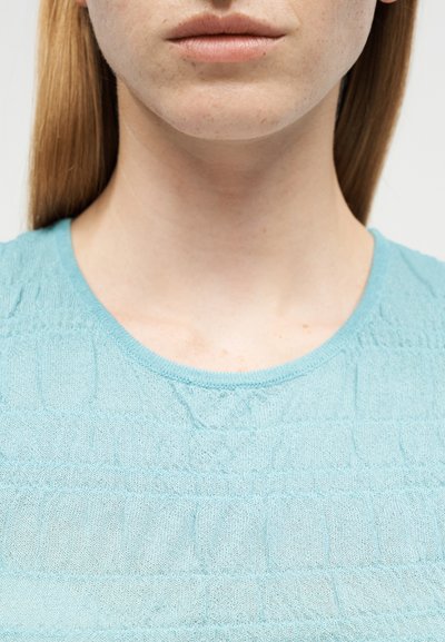 Κοντινό πλάνο του κάτω μέρους του προσώπου ενός ατόμου, του λαιμού και του άνω στήθους που φοράει ένα ανοιχτό μπλε υφασμάτινο πουλόβερ με ίσια κόκκινα μαλλιά.