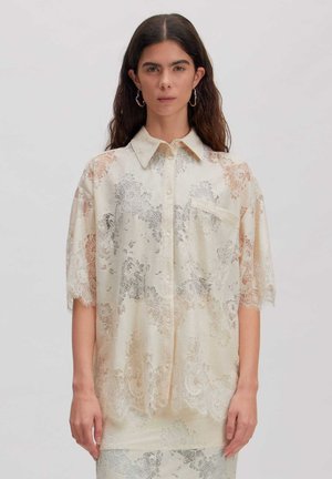 Frau mit dunklen Haaren trägt ein lockeres, cremefarbenes Spitzenhemd mit Blumenmustern, kurzen Ärmeln und einem Kragen vor einem schlichten Hintergrund.