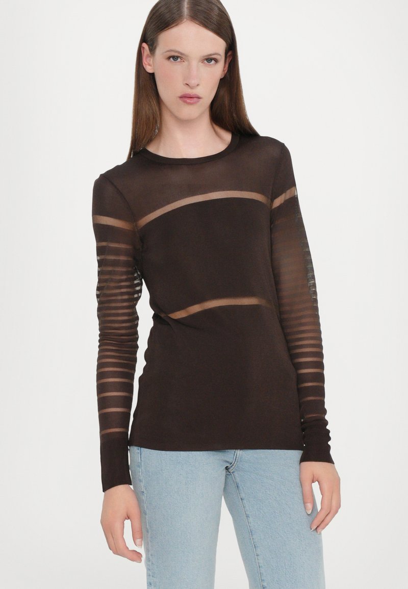 Maglietta marrone a maniche lunghe con strisce orizzontali trasparenti sulle maniche, caratterizzata da un colletto rotondo e una texture morbida. Abbinata a jeans azzurri chiari.