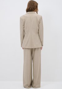 Beige räätälöity bleiseri, jossa on pitkät hihat ja kolmonen nappia, yhdistettynä leveälahkeisiin beigeihin housuihin. Silkkinen pinta, minimalistinen muotoilu.